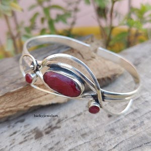 Peut inclure: Un bracelet manchette en argent avec trois pierres rouges. Le bracelet présente une grosse pierre ovale au centre et deux petites pierres rondes. Le design a une forme organique et fluide, avec la marque "luckyjewelart" visible.