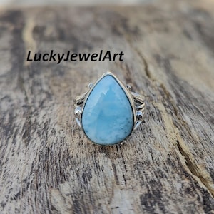 May include: A sterling silver ring featuring a pear-shaped, light blue larimar gemstone. The ring has an ornate, detailed setting with decorative elements on the sides. The text "LuckyJewelArt" is in the upper left corner.