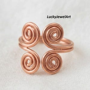 May include: A copper-coloured wire ring featuring an open design with four symmetrical spiral motifs. The metal wire is coiled into circular shapes at each end of the band. The text LuckyJewelArt is visible in the upper right corner of the image.