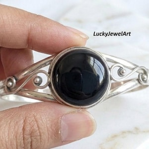 Könnte beinhalten: Ein silbernes Armband mit einem großen, runden, polierten schwarzen Stein in der Mitte. Das Metallband zeigt dekorative, geschwungene Drahtdetails auf beiden Seiten des Steins. Der Text LuckyJewelArt ist in der oberen rechten Ecke zu sehen.