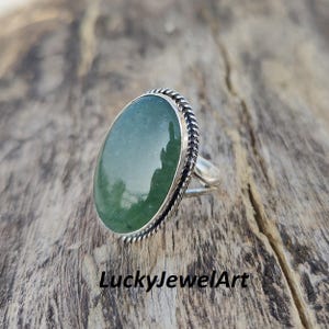 Könnte beinhalten: Ein Silberring mit einem großen, ovalen grünen Edelstein. Der Edelstein ist in einem Silberband mit einem geflochtenen Seildesign am Rand gefasst. Der Ring wird auf einer Holzoberfläche präsentiert. Der Text "LuckyJewelArt" ist sichtbar.