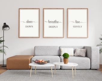 May include: Three framed wall art prints with the words "slow down", "breathe deeply", and "live gently" in black script. The prints are in light wood frames and hang above a gray sofa. A modern living room setting.