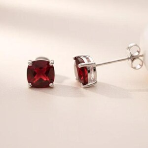 May include: A pair of silver stud earrings with square, deep red gemstones. The earrings are set in a four-prong setting, and the gemstones have a faceted appearance. The earrings are displayed on a light beige surface.