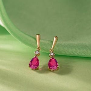 Peut inclure: Une paire de boucles d'oreilles pendantes dorées avec des pierres précieuses roses vives en forme de goutte. Chaque boucle d'oreille comporte une petite pierre scintillante au-dessus de la gemme rose. Les boucles d'oreilles sont présentées sur un fond vert doux.