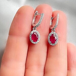 May include: A pair of silver hoop earrings featuring oval, deep red gemstones. Each gemstone is encircled by a halo of small, clear crystals. The earrings are shown against a soft, neutral background.