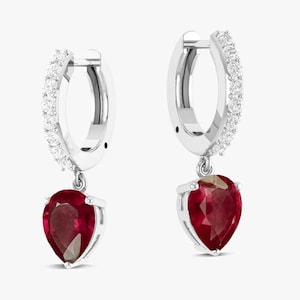 May include: A pair of silver hoop earrings with a row of small, clear stones along the top curve. Each earring has a pear-shaped, deep red gemstone suspended from the bottom.