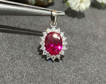 May include: A silver pendant with an oval, deep red gemstone at its center, surrounded by a halo of smaller, clear stones. The pendant is held by a pair of tweezers against a dark gray background, with white flowers in the background.