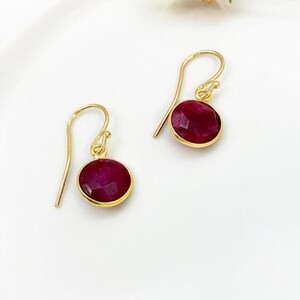 May include: A pair of gold drop earrings featuring round, deep red gemstones. The gemstones are set in a gold bezel, and the earrings have a simple hook design. The earrings are presented on a white surface.
