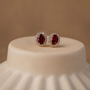 Op de afbeelding: Een paar goudkleurige oorstekers. Elke oorbel heeft een grote, ovale, dieprode edelsteen omringd door een halo van kleinere, heldere edelstenen. De oorbellen worden gepresenteerd op een wit, afgerond oppervlak tegen een neutrale achtergrond.