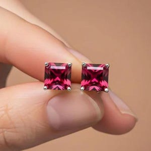Könnte beinhalten: Ein Paar quadratisch geschliffene Ohrstecker mit rubinroten Edelsteinen. Die Ohrringe sind in einem silberfarbenen Metall gefasst. Die Edelsteine haben ein facettiertes Design, das Licht reflektiert.