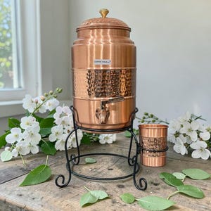 May include: A copper water dispenser with a hammered texture, a tap, and a lid with a gold-coloured knob. The dispenser sits on a black metal stand, with a matching copper cup. The text "COPPER YOGA" is visible on the dispenser.