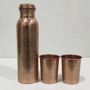 May include: A polished copper water bottle with a matching cap and two matching copper tumblers. The bottle and tumblers feature an intricate, embossed floral pattern. The text "COPPER YOGA" is printed on the upper section of the bottle.