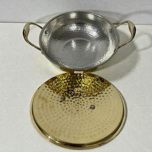 Op de afbeelding: Een gehamerde metalen kookpot met bijpassende deksel. De pot heeft twee goudkleurige handvatten en een zilveren binnenkant. De deksel is goudkleurig met een gehamerde textuur. De pot en deksel staan op een wit oppervlak.