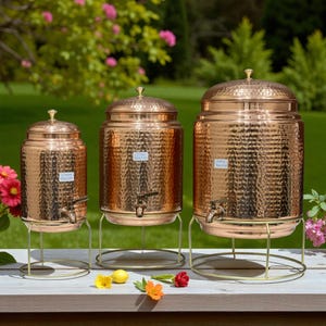 May include: Three copper beverage dispensers with a hammered texture, each with a spigot and lid. They are on gold-coloured metal stands. The dispensers are of varying sizes, arranged on a wooden surface with flowers in the foreground and a green background.