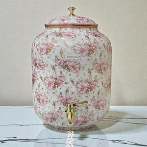May include: A white ceramic water dispenser with a floral pattern in pink and gold. The dispenser has a gold-colored spigot and a matching lid with a gold finial. The text "COPPER YOGA" is visible on the side.