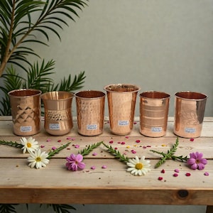 May include: A collection of six copper drinking glasses with different textures and patterns, including hammered and etched designs. Each glass has a small label that reads "Copper Royalty". The glasses are displayed on a wooden surface, decorated with flowers.