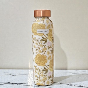 May include: A copper-colored water bottle with a floral design in yellow and white. The bottle has a copper-colored lid and the text "COPPER YOGA.in" on the front. The bottle is cylindrical and stands upright.