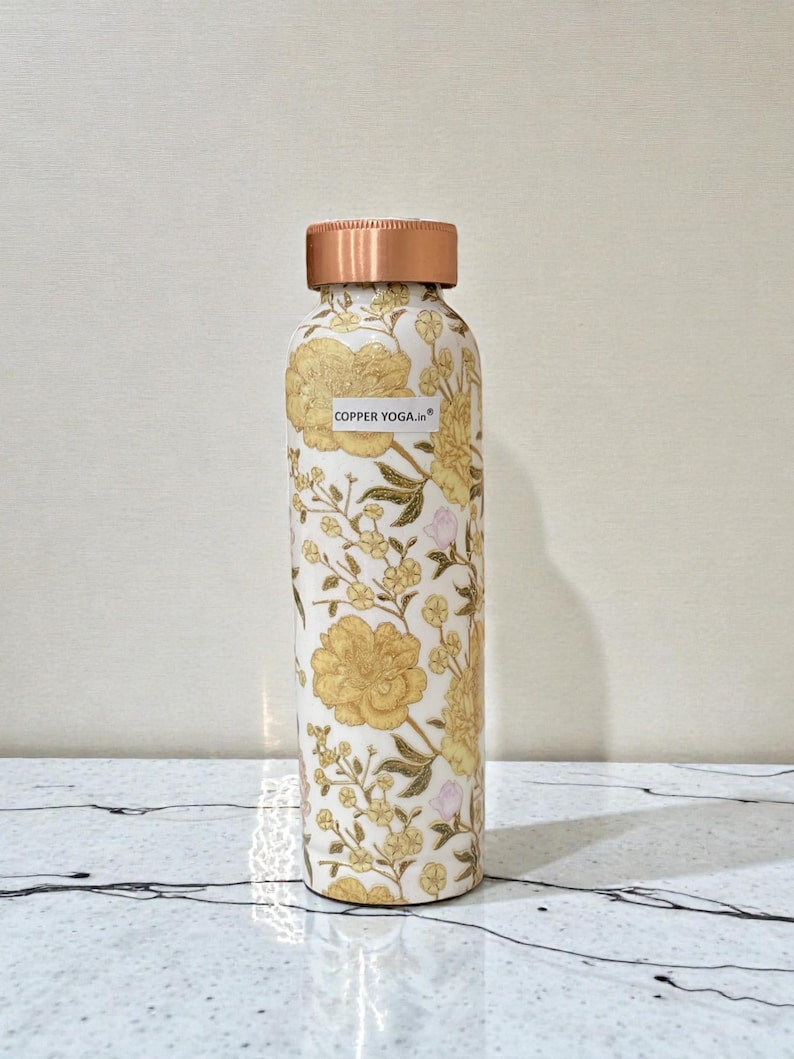 May include: A copper-colored water bottle with a floral design in yellow and white. The bottle has a copper-colored lid and the text "COPPER YOGA.in" on the front. The bottle is cylindrical and stands upright.