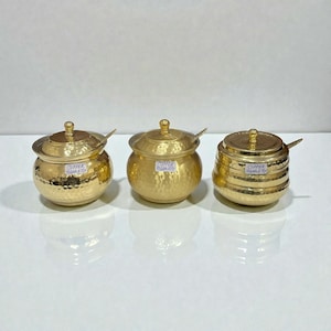 Puede incluir: Tres recipientes metálicos dorados con acabado martillado, tapas y pequeñas cucharas. Los recipientes son de diferentes formas y tamaños. Cada uno tiene una pequeña etiqueta que dice "COPPER GALLERY".