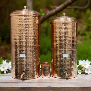 Può includere: Due contenitori per filtri d'acqua in rame con texture martellate, una piccola tazza in rame e un coperchio in rame. I contenitori hanno un rubinetto e una piccola etichetta con la scritta "COPPER SAFETY". Gli oggetti sono su una superficie di legno.