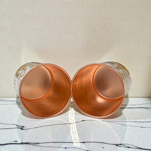 May include: Two copper-colored cylindrical glasses with a gold floral pattern on the exterior. The interior of the glasses has a shiny copper finish. The glasses are displayed on a white surface with black veining.
