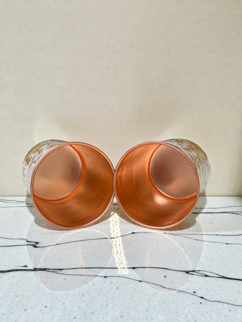 May include: Two copper-colored cylindrical glasses with a gold floral pattern on the exterior. The interior of the glasses has a shiny copper finish. The glasses are displayed on a white surface with black veining.