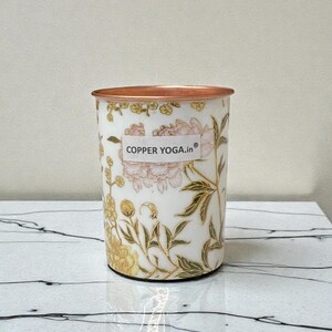 May include: A white tumbler with a copper rim and floral design. The tumbler features a pattern of pink and yellow flowers with green leaves. The text "COPPER YOGA.in" is printed on the front.