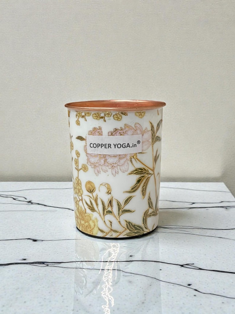 May include: A white tumbler with a copper rim and floral design. The tumbler features a pattern of pink and yellow flowers with green leaves. The text "COPPER YOGA.in" is printed on the front.