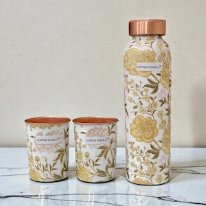 May include: A copper water bottle and two matching cups. The set features a floral design in gold and pink on a white background. The bottle and cups have copper rims and the text "COPPER YOGA.in".