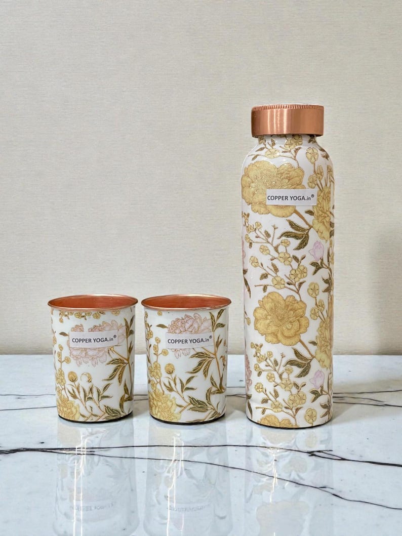 May include: A copper water bottle and two matching cups. The set features a floral design in gold and pink on a white background. The bottle and cups have copper rims and the text "COPPER YOGA.in".