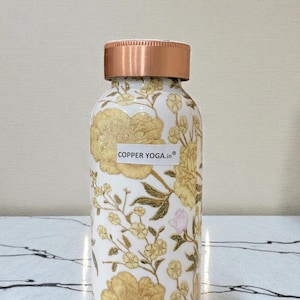 May include: A white water bottle with a copper lid and floral design. The bottle is covered in yellow flowers and green leaves. The text "COPPER YOGA.in" is on a white sticker on the bottle.