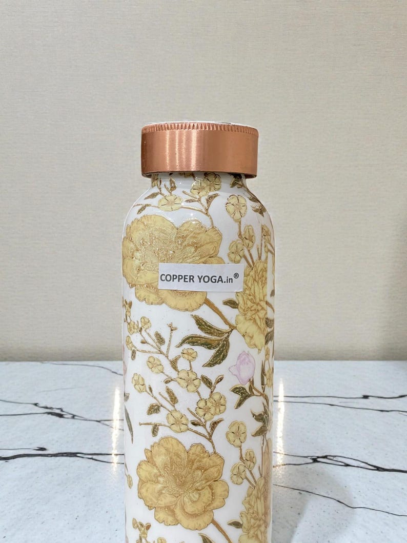 May include: A white water bottle with a copper lid and floral design. The bottle is covered in yellow flowers and green leaves. The text "COPPER YOGA.in" is on a white sticker on the bottle.