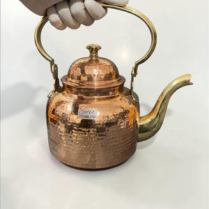 Puede incluir: Una tetera de cobre con acabado martillado, con asa y boquilla de latón. La tapa está rematada con un pequeño pomo de latón. La tetera está sujeta por una mano enguantada. El texto "COPPER YOGAA.ETS" es visible.