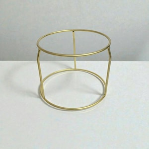 Puede incluir: Un soporte para plantas de metal dorado con una parte superior e inferior circulares, conectadas por soportes verticales. El soporte tiene un diseño minimalista y es adecuado para exhibir plantas o artículos decorativos. El soporte es de metal y tiene un acabado brillante.