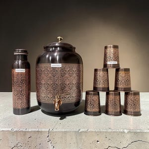 May include: A copper-toned water set featuring a large, round water dispenser with a spigot, a water bottle, and six matching cups. All pieces have a dark brown base with an ornate copper-colored floral pattern. The water bottle and dispenser have the text "Copper Yoga".