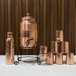 Puede incluir: Un juego de dispensador de agua de cobre con textura martillada, que incluye una botella, un dispensador grande sobre un soporte negro y varias tazas a juego. El dispensador tiene un grifo y una tapa.