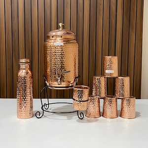 May include: A copper water dispenser set with a hammered texture. The set includes a large dispenser with a spigot, a water bottle, and several small drinking cups. The dispenser sits on a black metal stand. The background is a wooden wall.
