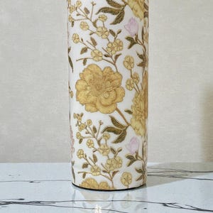 May include: A cylindrical water bottle with a floral design. The bottle features large yellow flowers, smaller yellow blossoms, and pink buds on a white background. The text "COPPER YOGA.in" is visible at the top.
