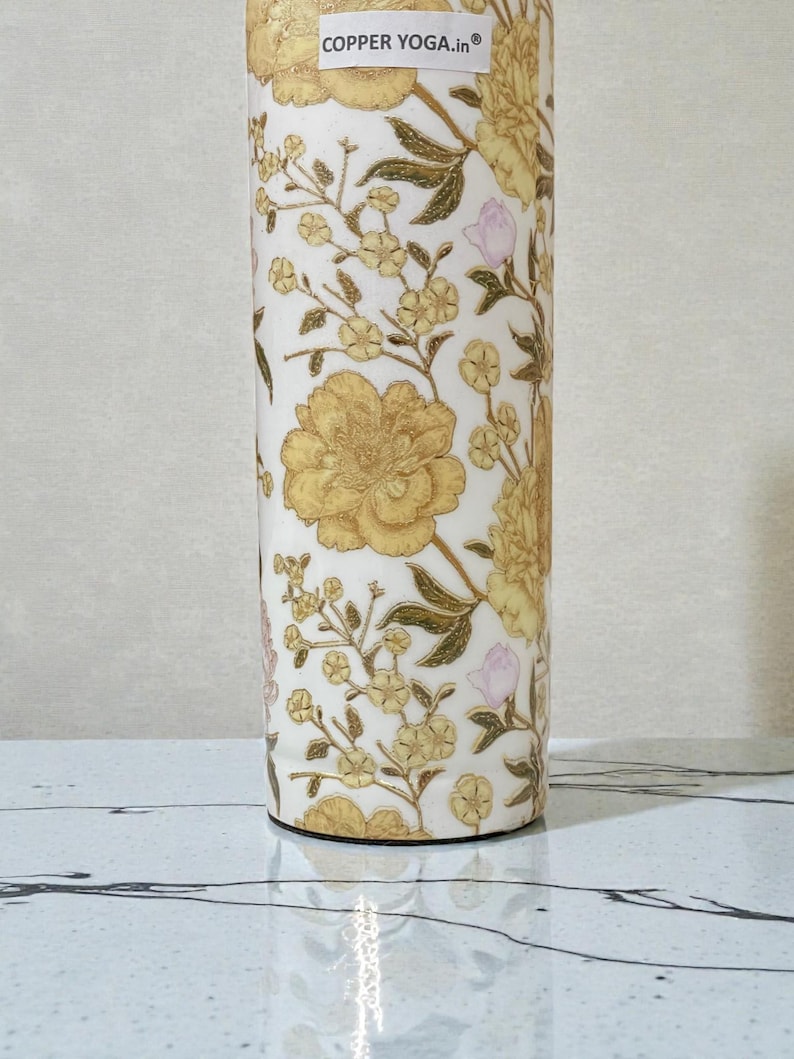 May include: A cylindrical water bottle with a floral design. The bottle features large yellow flowers, smaller yellow blossoms, and pink buds on a white background. The text "COPPER YOGA.in" is visible at the top.