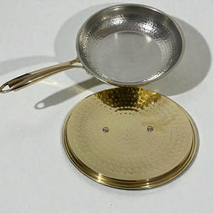 Puede incluir: Una sartén de acero inoxidable con un mango dorado y una tapa a juego. La sartén y la tapa tienen una textura martillada. La tapa está a un lado, revelando el interior de la sartén.