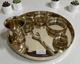 Puede incluir: Un juego de thali de color latón con varios cuencos pequeños, una taza alta y una cuchara. El juego está sobre una bandeja redonda. El texto "COPPER YOGAA.ETSY" es visible en la bandeja. Pequeñas suculentas en macetas están en el fondo.