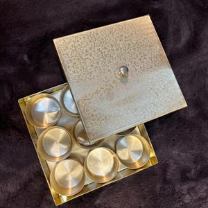 May include: A gold-coloured spice box with a floral pattern on the lid. The box is open, revealing several small, round compartments. The box has a small, round knob on the lid. The box is on a dark brown surface.