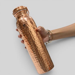 May include: A cylindrical copper water bottle with a hammered texture and a metallic finish. The bottle features a screw-top lid and a small black logo printed on the upper body that reads Copper Yoga with a tree icon above the text.