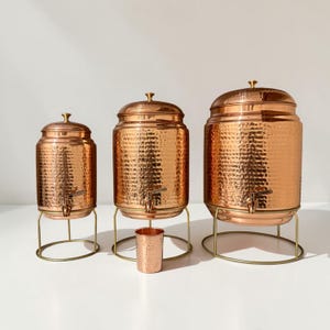 Peut inclure: Trois distributeurs de boissons en cuivre martelé de tailles différentes, chacun doté d'un couvercle assorti, d'un robinet en métal et d'un support circulaire en fil métallique. Une petite tasse en cuivre martelé est placée devant le distributeur central.