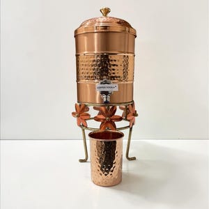 May include: A copper water dispenser with a hammered texture, a lid, and a spigot. The dispenser sits on a decorative stand with orange flower accents. A matching copper cup is in front of the dispenser. The text "COPPER YOGA" is visible.