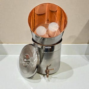 Puede incluir: Un filtro de agua de cobre y acero inoxidable con tapa y grifo. La sección superior es de color cobre y el cuerpo principal es de acero inoxidable. Dos objetos blancos están dentro de la sección de cobre.