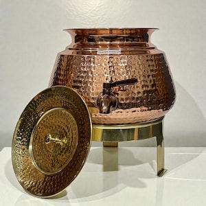 Puede incluir: Un dispensador de agua de cobre martillado con grifo, tapa y soporte de latón. El dispensador tiene un cuerpo ancho y redondeado y una parte superior acampanada. La tapa es redonda con una textura martillada. El dispensador está etiquetado "COPPER YOGA".