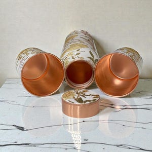 May include: A set of copper-colored metal containers with a floral design in gold and white. The set includes a tall, cylindrical container, two cups, and a small, round container with a lid. The interiors of the containers are a shiny copper color.