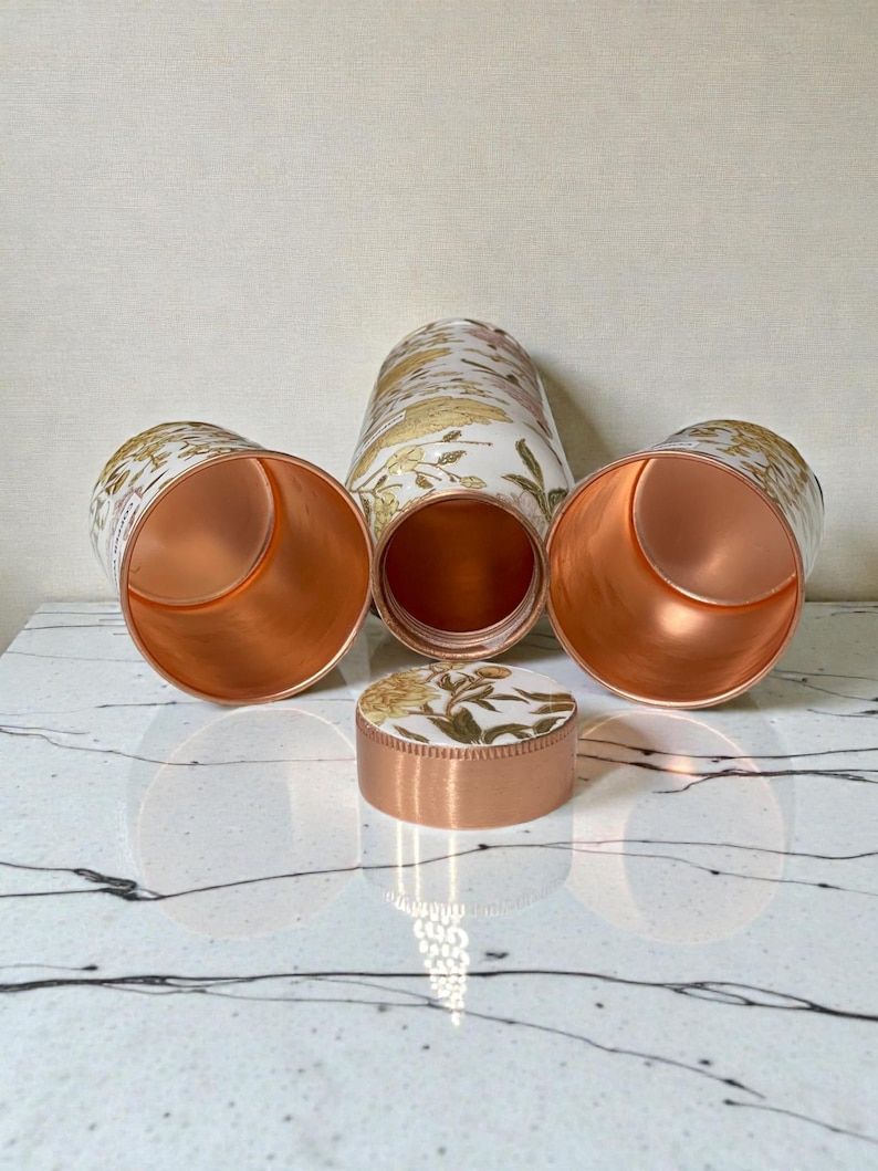 May include: A set of copper-colored metal containers with a floral design in gold and white. The set includes a tall, cylindrical container, two cups, and a small, round container with a lid. The interiors of the containers are a shiny copper color.