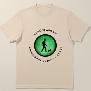 Könnte beinhalten: Ein naturfarbenes T-Shirt mit einem kreisförmigen Grafikdesign, das eine Person mit einem Hund zeigt, umgeben von dem Text "Crossing with my emotional support chaos". Das Design ist in Schwarz und Grün gehalten.